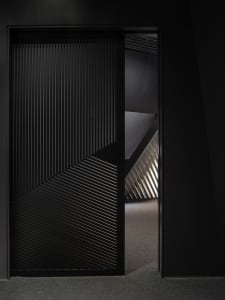 Modern black sliding door with diagonal metal slats leading to a staircase with similar design.