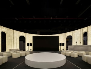 A minimalist conference room with a round, white platform at the center, surrounded by upholstered seating and concrete walls