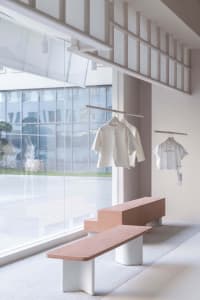 A minimalist retail store with concrete and wood elements, featuring clothing racks and seating areas.