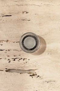 A minimalist glass coaster with a dark circular design on a textured, beige stone surface.