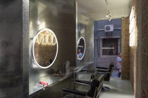 A minimalist salon with metallic and brick textures, featuring round mirrors with neon lights.