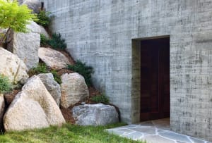 A modern entrance made of concrete with a large stone door and landscaping.