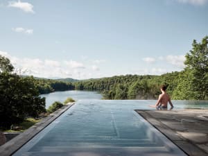 Modern residential architecture featuring a glass infinity pool overlooking a scenic lake and forested landscape.