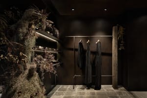 A modern clothing store with dark decor, featuring a wooden pillar, shelves with plants, and clothing on a hanging rail.