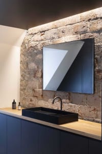 A modern bathroom featuring a black sink and faucet on a wooden counter against a brick wall with a skylight.