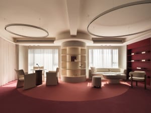 A modern interior space with a circular table, chairs, and bookshelf, featuring neutral-toned furniture and a red carpet.