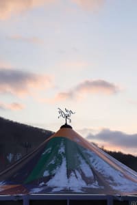 A colorful, triangular tent covered in snow with a weather vane on top.