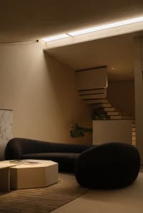 A minimalist living room with a black sofa, a round wooden coffee table, and concrete walls and floor.