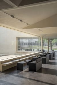 A spacious, modern cafe interior featuring concrete walls, high ceilings, and large windows overlooking a green courtyard.