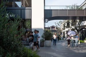 People walking on a walkway surrounded by retail stores with concrete and glass facades, greenery, and a pedestrian bridge.