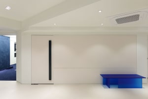 A minimalist lobby featuring a glass and metal table with a blue top on a white floor and wall