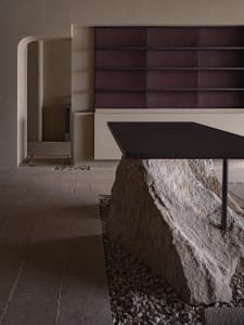 A modern bench featuring a unique design with a rock base, placed in a minimalist space with a concrete wall and floor.