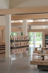 A minimalist modern library with wooden bookshelves, concrete flooring, and large windows allowing natural light.