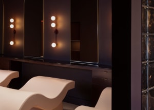 A modern beauty salon features a row of illuminated mirrors, glass partitions, and sleek seating areas.