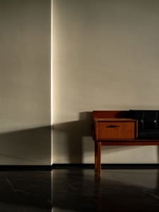 A minimalist interior with a wooden table and black leather seating against a plain wall and marble floor.