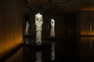 An interior image showcasing a glass exhibit space with illuminated mannequins and dresses.