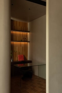 A modern minimalist office with glass desk, red book, black chair, plant, and shelving with books and decorative items.