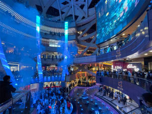 A bustling shopping mall interior with modern design elements including glass panels, LED lighting, and concrete surfaces.