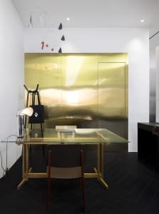 A minimalist office with a transparent glass desk, wooden chair, and metallic accents.