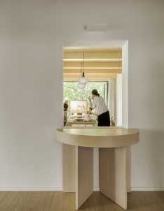 A minimalist home office featuring a wooden desk with an open storage in the center, a chair, and a view of a room with a pai
