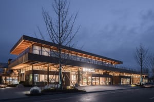 A modern shopping center exterior illuminated at night with wooden structures, glass walls, and concrete elements.