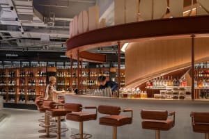 A modern retail bar with a curved wooden counter, leather stools, and a variety of bottles on display.