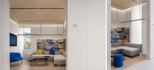 A modern office lounge area with white and concrete furniture, featuring a large grey sofa, blue chairs, and a blue pouf.