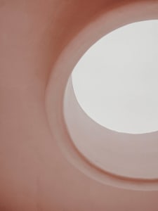 View of a minimalist architectural space with light-colored concrete walls and a circular opening allowing natural light to e