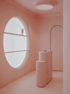 A minimalist interior space with concrete and plaster surfaces, featuring a large oval window, rounded shelves, and smooth, c