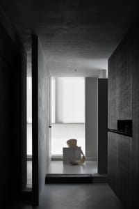 A minimalist interior space with concrete walls and floor, featuring a modern sculpture on a platform in a hallway.