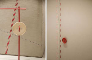 A display featuring a large beige button with a red rod and crosshatch lines drawn on a fabric surface, with a smaller red bu