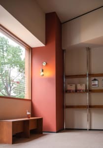 A minimalist coffee shop interior with a window displaying greenery, a warm light fixture, a wooden bench with a beverage cup