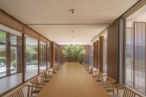 A minimalist cafeteria featuring wooden and glass elements, with a long wooden table and chairs.