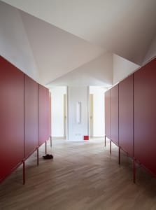 A minimalist hallway with red cabinets on either side and a light-colored wooden floor. There is a red object on the floor.