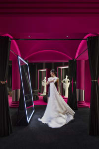 A woman wearing a long white dress in a modern retail space with pink lighting and black curtains.