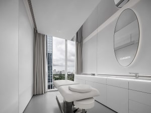 A modern medical office with white walls, a white medical bed, and a large window with a city view.