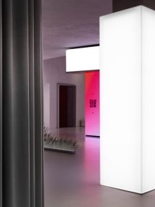 Modern minimalist hallway with a bright white lighted column and a pink-lit room in the distance.