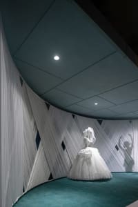A wedding dress displayed in a contemporary, minimalist space with white fabric and turquoise carpet.