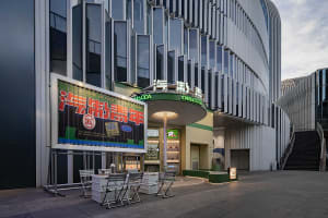 Modern commercial building exterior with metal cladding and large glass windows, featuring a large signboard and seating area