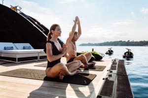 A couple practicing minimalist yoga on a boat deck near the water with a luxury yacht in the background.