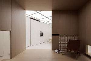 Modern minimalist residential interior with wooden walls and floor, a chair, and a view of the kitchen area.