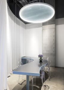 Luminous oval light fixture, white curtains, and glass chairs surround a sleek, modern conference room table.