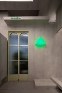 A minimalist retail interior with concrete walls and a green neon triangle light on a textured concrete wall next to a glass 
