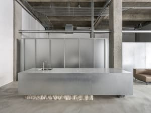 A modern kitchen with a large grey counter and industrial-style office space in the background, featuring exposed concrete wa