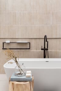 A minimalist bathroom with ceramic tiles and a concrete bathtub, featuring a wooden stool with a vase and a book.