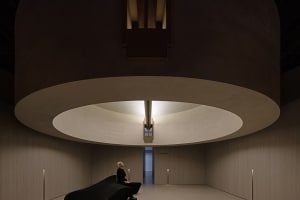A minimalist interior with a white concrete floor and ceiling, featuring a circular light fixture and a woman sitting on a bl