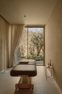 A minimalist spa treatment room featuring a concrete bed and floor, with a window overlooking a serene outdoor garden.