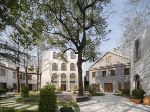 Modern building exterior featuring brown brick and white concrete materials, with a lush green courtyard and tall trees.