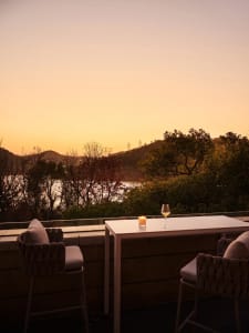 A cozy outdoor seating area with a wooden table and chairs, overlooking a scenic sunset and natural landscape.