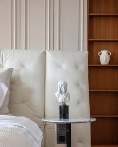A minimalist bedroom featuring a leather headboard with tufted cushions, a marble bedside table, and a wooden bookshelf with 